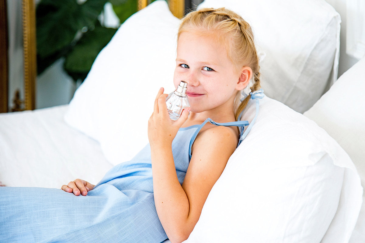 A smiling little girl in a blue dress smelling a perfume bottle