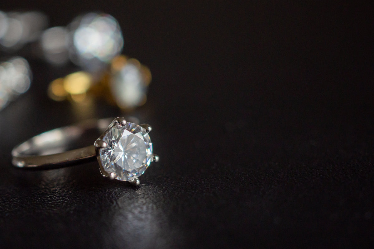 A sparkling solitaire ring resting on a black surface, with more rings softly out of focus behind it
