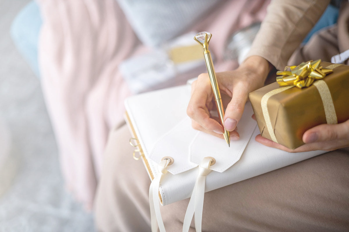 A hand writing a gift tag with a gold pen while holding a small box tied with gold ribbon